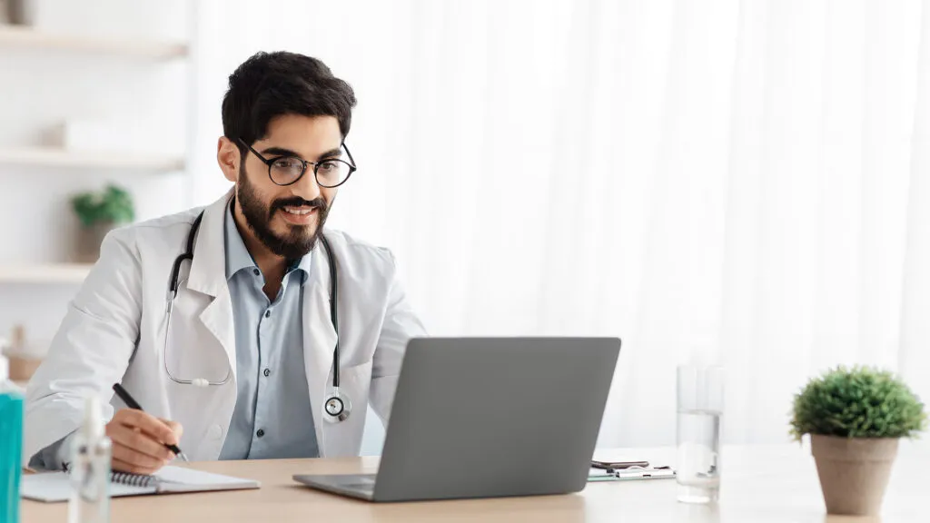 Profesional de salud frente a una computadora portátil, con un bolígrafo y un cuaderno